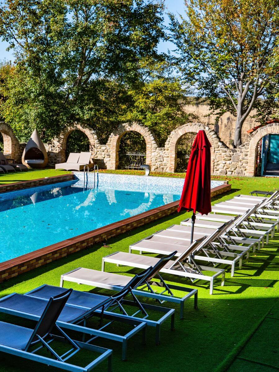 Sun loungers and red umbrellas around the pool, with a historic stone wall and autumn trees behind.