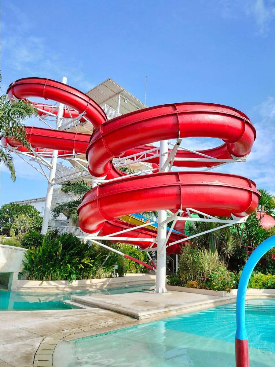 Twisting red water slides at a resort pool, surrounded by plants under a clear blue sky