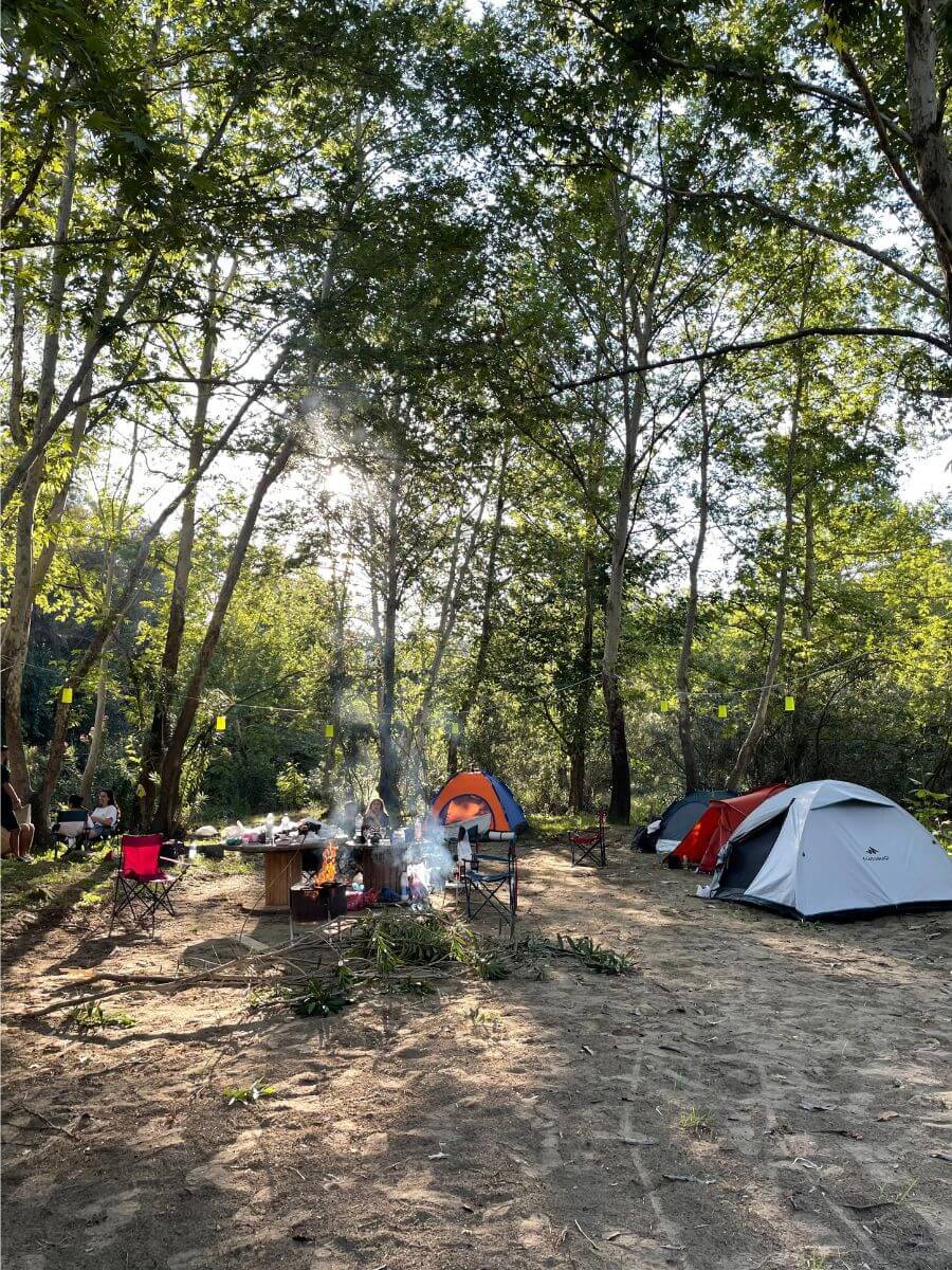 Gray, red and orange dome tents on sandy ground under tall trees, with a campfire, smoke and people